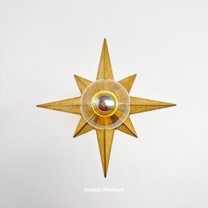 May include: A gold-toned star-shaped wall sconce featuring a central clear glass globe light fixture. The metal star has eight points of varying lengths and a smooth finish. The text Amanda Hardware is visible at the bottom of the image.