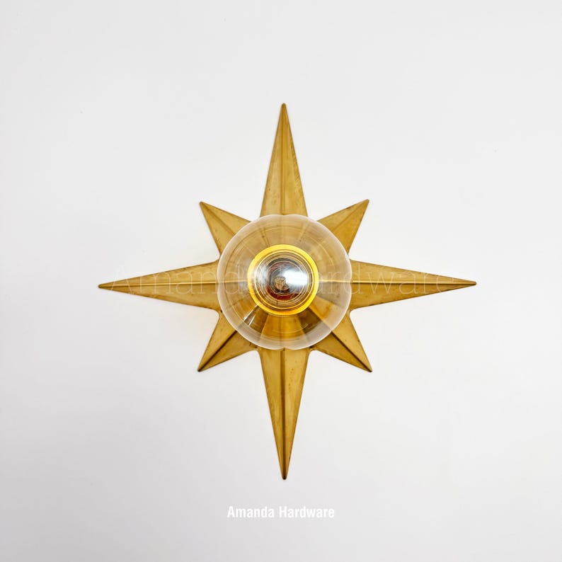 May include: A gold-toned star-shaped wall sconce featuring a central clear glass globe light fixture. The metal star has eight points of varying lengths and a smooth finish. The text Amanda Hardware is visible at the bottom of the image.