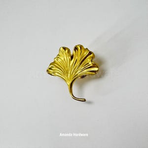 May include: A gold-colored metal ginkgo leaf-shaped door pull. The leaf is detailed with veins and has a curved stem.