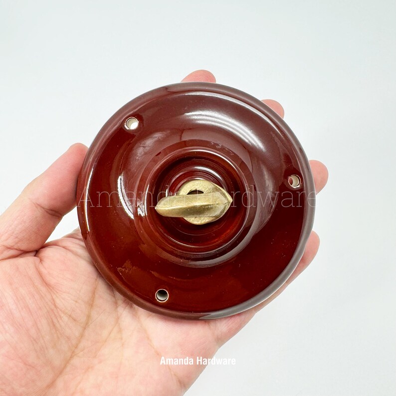 May include: A round, dark brown ceramic door knob with a brass-colored handle. The knob has a glossy finish and three small holes around the edge. The text "Amanda Hardware" is visible at the bottom.