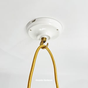 Puede incluir: Un dosel de techo de cerámica blanca con un gancho de latón y un cable eléctrico dorado cubierto de tela. El texto "Amanda Hardware" es visible en la parte inferior. Componente de luminaria.