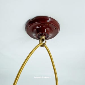 May include: A maroon ceiling canopy with a brass hook and a gold fabric-covered electrical cord. The canopy is mounted on a white ceiling. The cord is looped and ready for a light fixture.