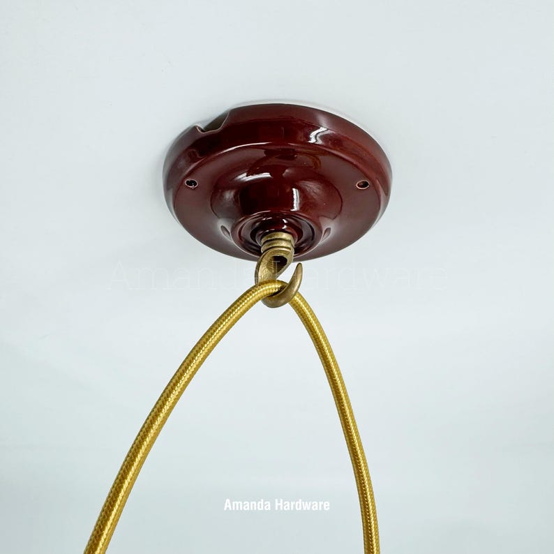 May include: A maroon ceiling canopy with a brass hook and a gold fabric-covered electrical cord. The canopy is mounted on a white ceiling. The cord is looped and ready for a light fixture.