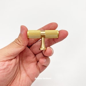 May include: A gold-toned T-shaped cabinet knob with a textured cylindrical handle. The knob is held in a hand.