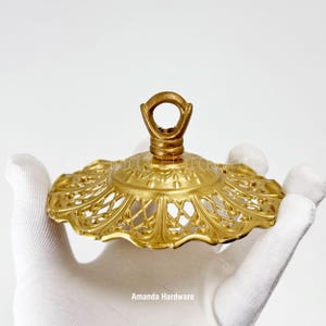 May include: A circular gold-toned metal ceiling canopy with an intricate filigree pattern along the scalloped edge and a central loop fixture at the top. The hardware features a textured finish and is branded with the text Amanda Hardware.