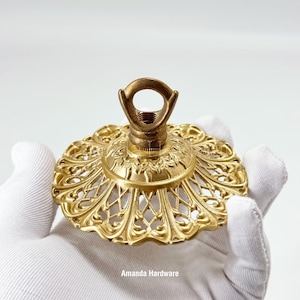 May include: A circular gold-toned metal ceiling canopy featuring an intricate openwork filigree pattern and a central threaded loop attachment. The hardware displays a textured finish with embossed floral details and the text Amanda Hardware.
