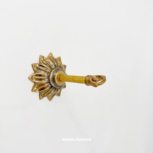 May include: A decorative curtain tieback in a sunflower design. The tieback is made of brass and features a sunflower-shaped base with detailed petals. The arm extends outward with a ring at the end for holding curtains. The text "Amanda Hardware" is at the bottom.
