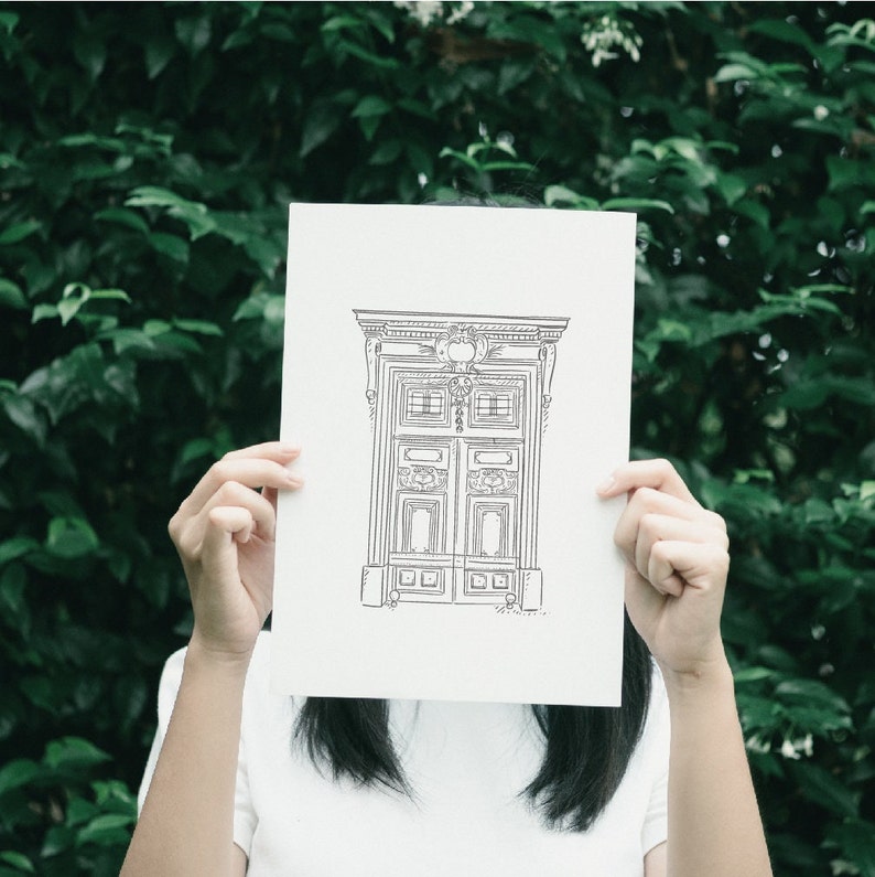 A black and white line drawing of a door with ornate details. The door is framed by a rectangular archway with a decorative top. The door has two panels with a decorative design and a small window in each panel. The door is set against a white background.