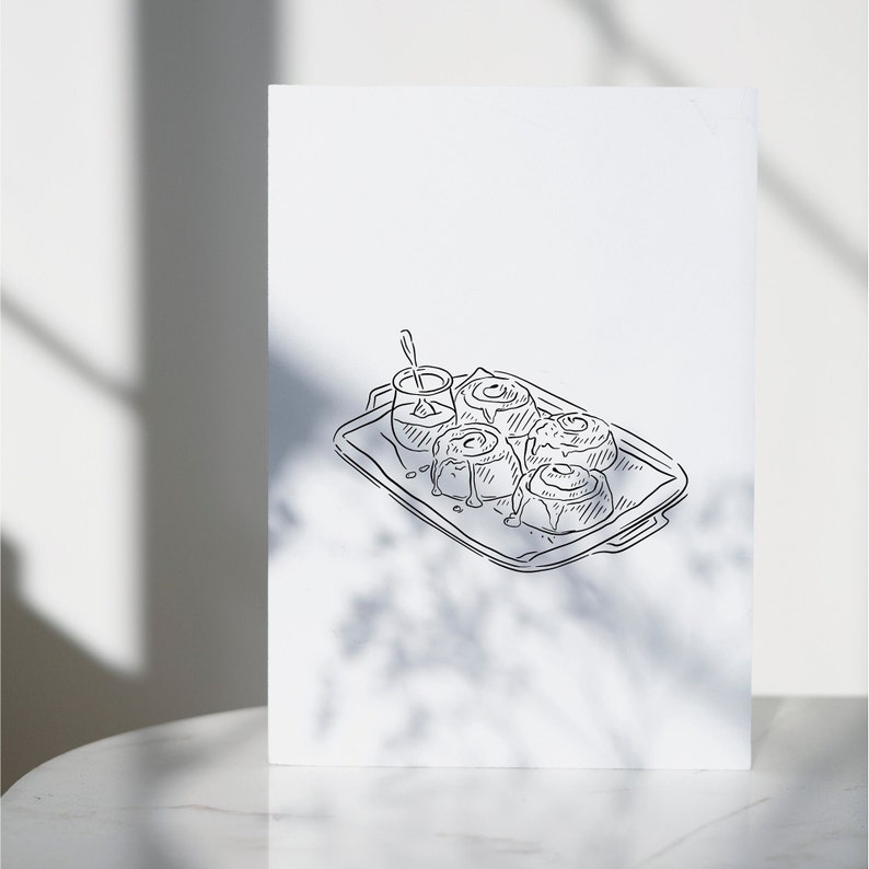 A black and white line drawing of a tray of cinnamon rolls with a small dish of icing on the side.