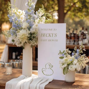 May include: A white rectangular sign with the text WELCOME TO BECKY'S BABY SHOWER and a green line drawing of a rubber duck. The sign is flanked by two white vases containing arrangements of light blue and white flowers on a wooden surface.