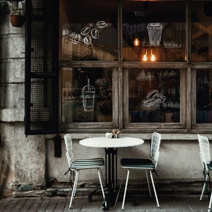 A white table with two white chairs sits in front of a shop window. The window has a wooden frame and is decorated with drawings of coffee beans and cups.