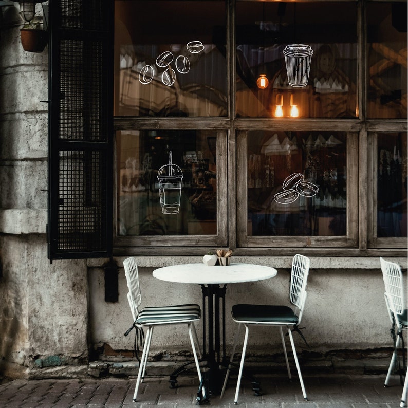 A white table with two white chairs sits in front of a shop window. The window has a wooden frame and is decorated with drawings of coffee beans and cups.