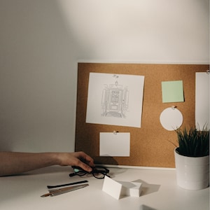 A corkboard with a white frame and a white background. The corkboard has a drawing of a doorway with a light above it, a green sticky note, a white circle, and a white note card. A white note card is also on the table below the corkboard. There are also a pair of black glasses, a silver ruler, and a black pen on the table. A potted plant with green leaves is on the table to the right of the corkboard.