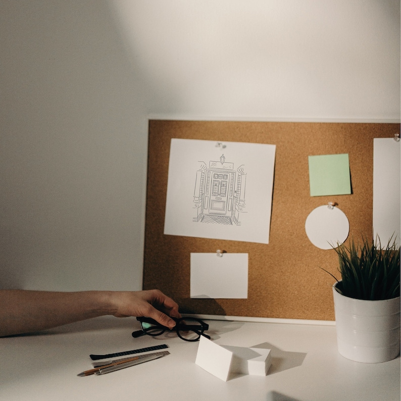 A corkboard with a white frame and a white background. The corkboard has a drawing of a doorway with a light above it, a green sticky note, a white circle, and a white note card. A white note card is also on the table below the corkboard. There are also a pair of black glasses, a silver ruler, and a black pen on the table. A potted plant with green leaves is on the table to the right of the corkboard.
