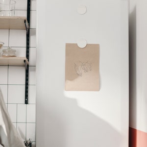 Puede incluir: Un dibujo lineal simple de dos manos que se sostienen, impreso en una tarjeta de papel marr&oacute;n y fijado a un refrigerador blanco con imanes.