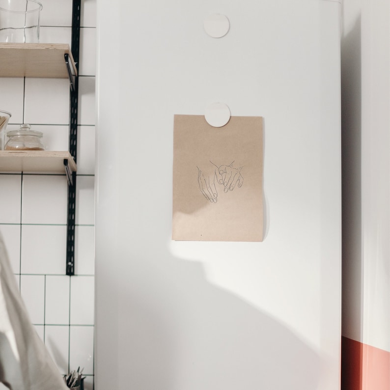 Puede incluir: Un dibujo lineal simple de dos manos que se sostienen, impreso en una tarjeta de papel marr&oacute;n y fijado a un refrigerador blanco con imanes.