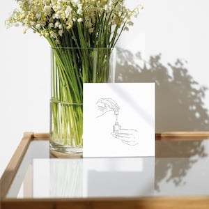Puede incluir: Una tarjeta blanca con un dibujo en blanco y negro de una mano sosteniendo una botella y un gotero. La tarjeta est&aacute; sobre una mesa de madera con un jarr&oacute;n de flores blancas en el fondo.