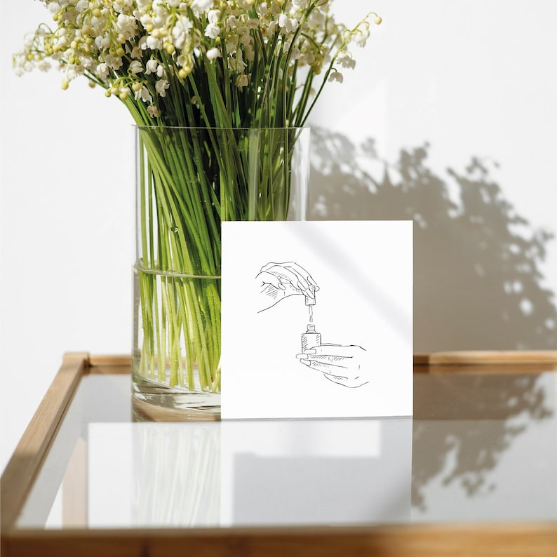 Puede incluir: Una tarjeta blanca con un dibujo en blanco y negro de una mano sosteniendo una botella y un gotero. La tarjeta est&aacute; sobre una mesa de madera con un jarr&oacute;n de flores blancas en el fondo.