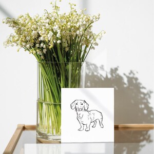 A black and white line drawing of a dachshund dog on a white card. The card is sitting on a wooden table next to a vase of white lily of the valley flowers.