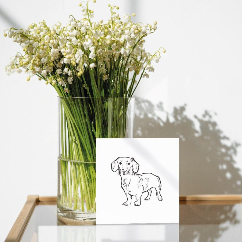 A black and white line drawing of a dachshund dog on a white card. The card is sitting on a wooden table next to a vase of white lily of the valley flowers.
