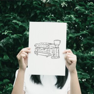 A black and white line drawing of a coffee machine on a white piece of paper held by a person.