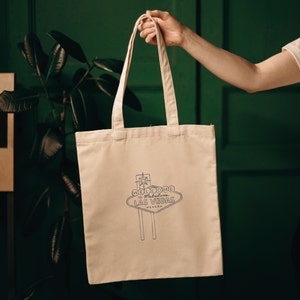 A white canvas tote bag featuring a black and white graphic of the "Welcome to Fabulous Las Vegas" sign. The bag has black straps.