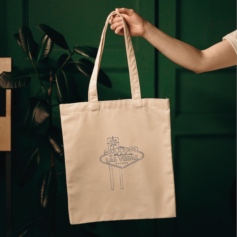 A white canvas tote bag featuring a black and white graphic of the "Welcome to Fabulous Las Vegas" sign. The bag has black straps.