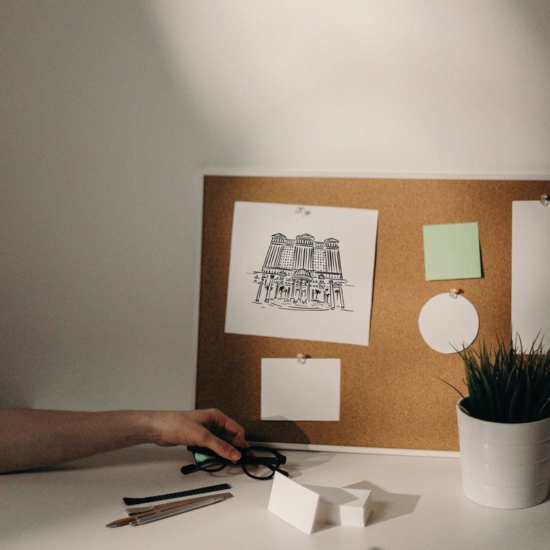 A corkboard with a black and white drawing of a building, a green sticky note, a white circle, and a white note. A potted plant with green leaves sits on a white surface in front of the corkboard.