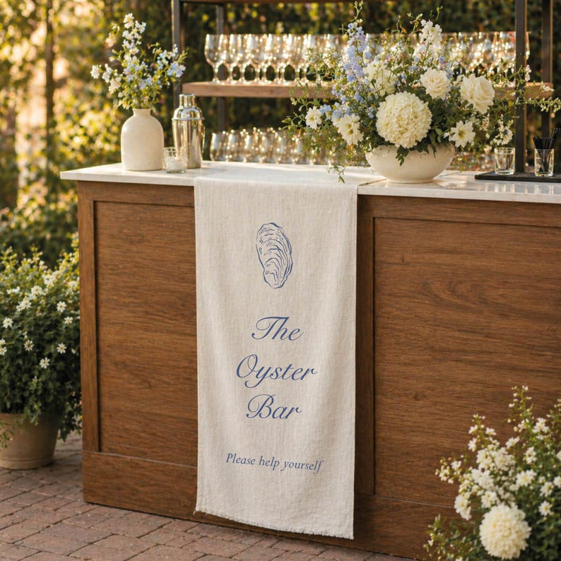 Puede incluir: Un bar de madera con encimera blanca y una pancarta de lino que dice "The Oyster Bar". La pancarta presenta una ilustraci&oacute;n de ostra azul. El bar est&aacute; decorado con arreglos florales y cristaler&iacute;a.