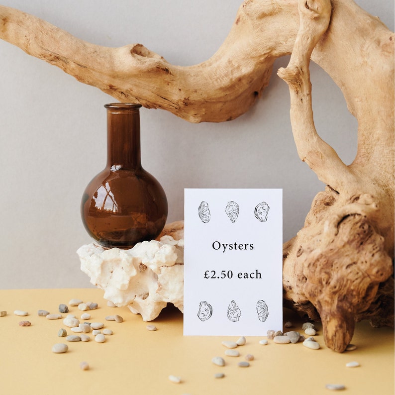 A white card with black text and illustrations of oysters. The card reads "Oysters £2.50 each". The card is sitting on a piece of white coral with a brown glass bottle behind it.
