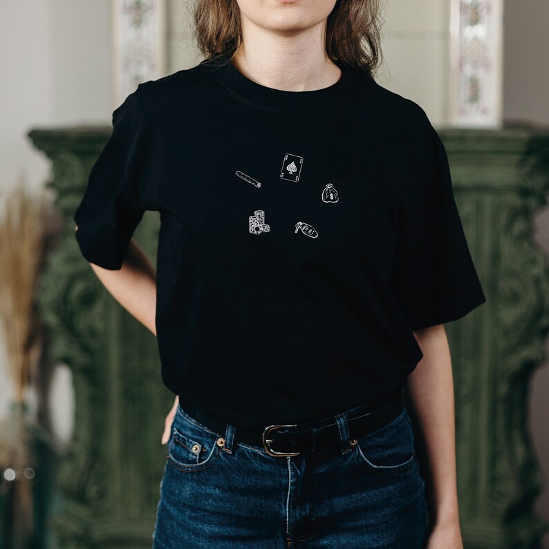A black t-shirt with a white graphic design of playing cards, a bag of money, a cigar, and dice.
