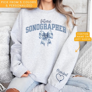 May include: Gray sweatshirt with the text "future sonographer" in blue with a blue floral bow. The left sleeve has a heart and the text "Maribel" in blue.