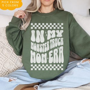 May include: Green sweatshirt with a white graphic that says "In My Monster Truck Mom Era" with a checkered pattern above and below the text.