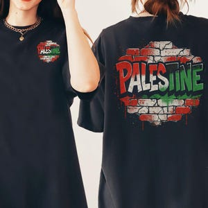 May include: Black t-shirt featuring the word "PALESTINE" in red and green, designed to resemble graffiti on a brick wall. The shirt has a smaller version of the design on the front.