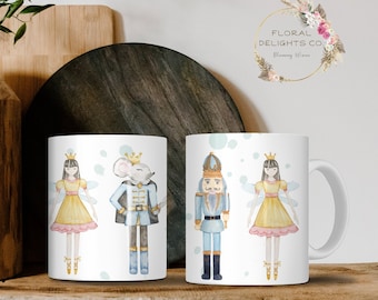 May include: Two white ceramic mugs with a Nutcracker and Sugar Plum Fairy theme. The mugs feature watercolor illustrations of a Nutcracker soldier, a Sugar Plum Fairy, and a mouse king. The mugs are on a wooden shelf.