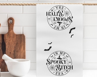 May include: White kitchen towel with a black and white graphic design that says "In My Spooky Bitch Era" with a witch's hat, spider web, and bats.