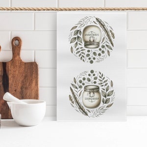 Puede incluir: Dos toallas de cocina blancas con una ilustración verde y marrón de un frasco etiquetado "SOURDOUGH" rodeado de hojas y plumas.