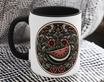 May include: White ceramic mug with a black rim and handle. The mug features a circular design of a watermelon slice surrounded by green leaves and red flowers.