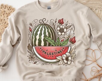 May include: A beige sweatshirt with a graphic design of a watermelon slice with the word "PALESTINE" on it. The watermelon is surrounded by flowers and leaves.