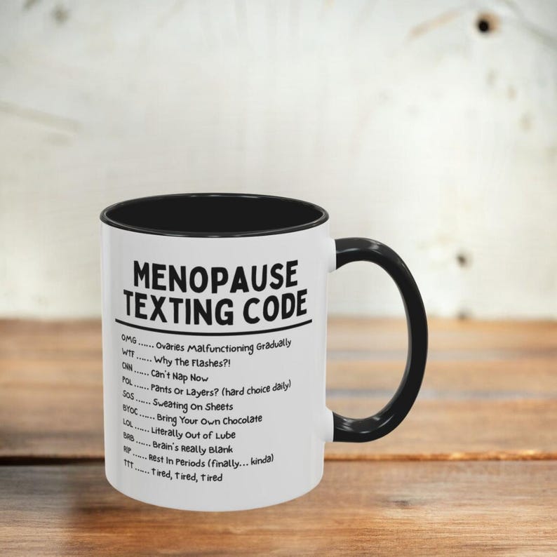 May include: White ceramic mug with a black interior and handle. The mug features the text "MENOPAUSE TEXTING CODE" with a list of acronyms and their meanings. The mug is on a wooden surface.