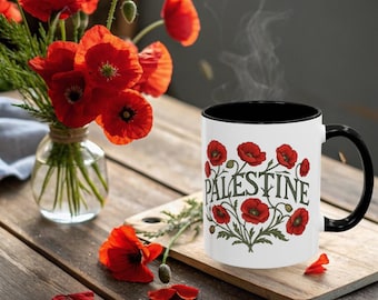 May include: A white coffee mug with a black interior and handle, featuring red poppy flowers and the word "PALESTINE" in green. A bouquet of red poppies in a glass vase and a wooden cutting board are also in the image.