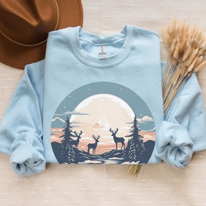 May include: A light blue sweatshirt with a graphic of a full moon, three deer, and trees in a forest setting. The graphic is in a circle with a dark blue outline.