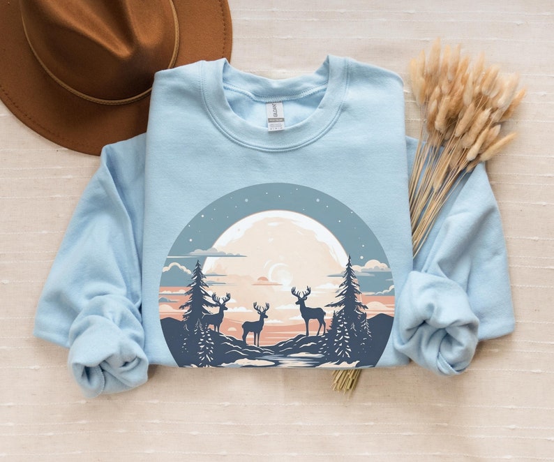 May include: A light blue sweatshirt with a graphic of a full moon, three deer, and trees in a forest setting. The graphic is in a circle with a dark blue outline.