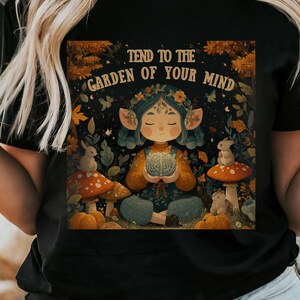 Puede incluir: Camiseta negra con la frase "TEND TO THE GARDEN OF YOUR MIND" y una ilustración fantástica. La obra de arte representa una figura sentada sosteniendo un cerebro, rodeada de setas, calabazas y elementos forestales. El diseño tiene un tema de fantasía.