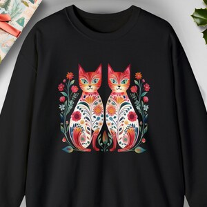 May include: Black sweatshirt with a colorful illustration of two cats with floral patterns. The cats are facing forward and have white eyes.