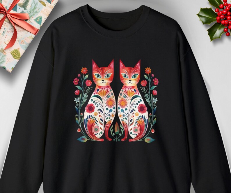 May include: Black sweatshirt with a colorful illustration of two cats with floral patterns. The cats are facing forward and have white eyes.