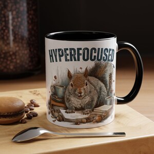 May include: A white ceramic mug with a black interior and handle, featuring the text "HYPERFOCUSED" and a detailed illustration of a squirrel surrounded by coffee cups, books, and nuts. The mug sits on a wooden surface.
