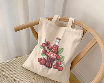 Könnte beinhalten: Eine weiße Canvas-Tragetasche mit einem Aufdruck einer roten Coca-Cola-Flasche und roten Kirschen. Die Flasche ist von grünen Blättern und weißen Blüten umgeben.