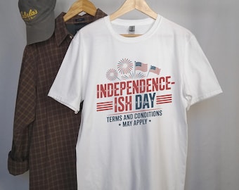 Puede incluir: Una camiseta blanca con el texto "INDEPENDENCE-ISH DAY" en rojo y azul, con fuegos artificiales y banderas americanas. Una camisa a cuadros marrón y una gorra de béisbol gris también cuelgan de una percha.