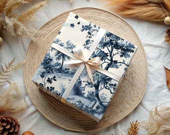 Könnte beinhalten: Eine weiße Geschenkbox mit einem blauen und weißen Blumenmuster. Die Box ist mit einem weißen Satinband gebunden und liegt auf einem geflochtenen Stroh-Untersetzer.